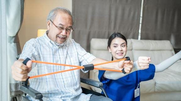 Person in a wheelchair doing resistance band exercises with assistance from a healthcare worker.