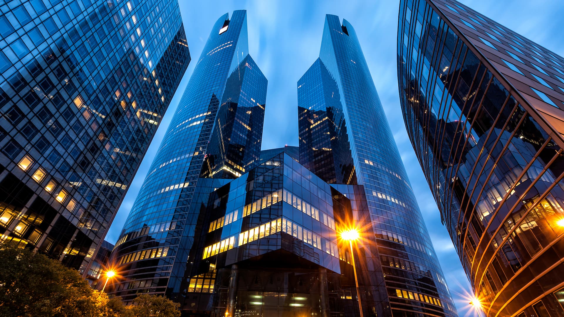 Modern glass skyscrapers viewed from street level at dusk, with illuminated office windows.