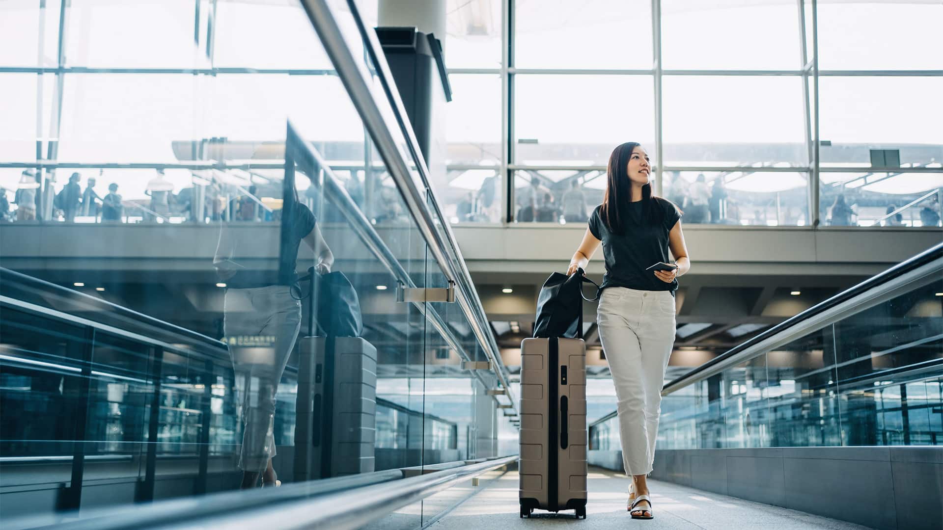 A women carrying a passport, purse, and suitcase while walking in an airport concourse