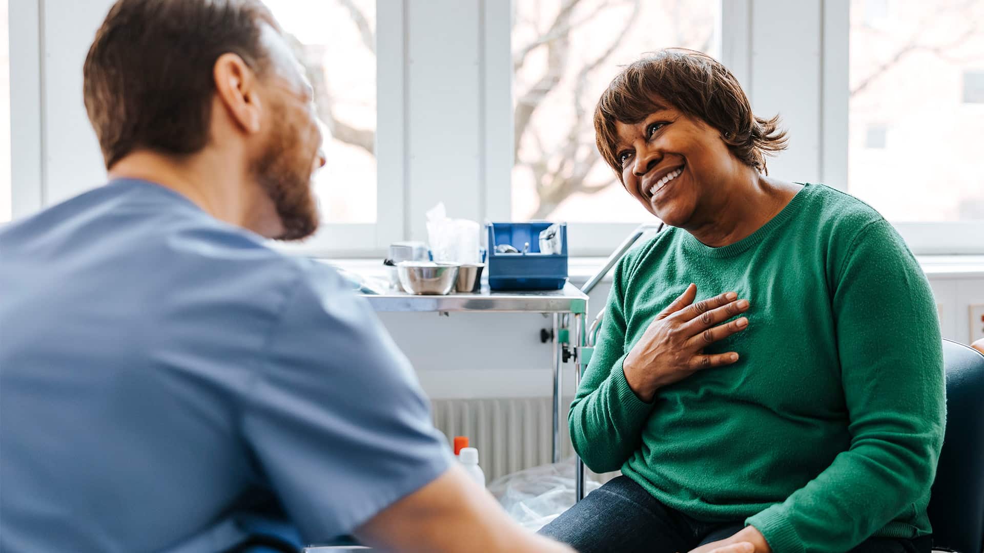 A happy senior woman with her hand on her chest talking with a male doctor while sitting in a medical examination room