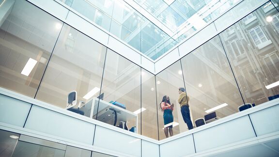 Two people standing and talking inside a bright, multi-story glass office atrium overlooking workspaces.