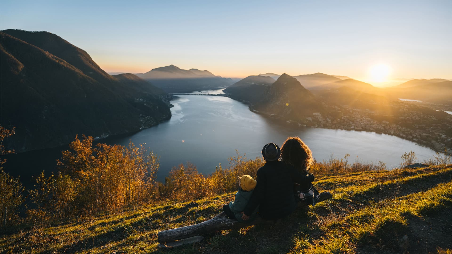 A family sitting on a mountain top looking out at the lake, sunset, and other mountains