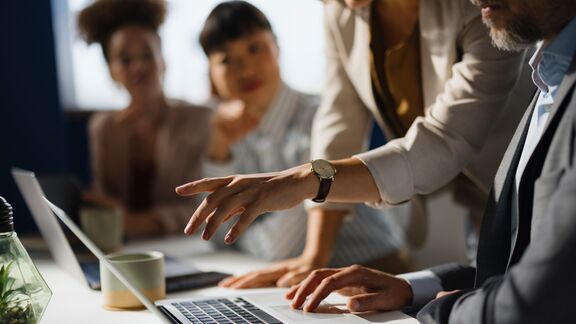Colleagues gathered around a laptop as one person points at the screen during a discussion.