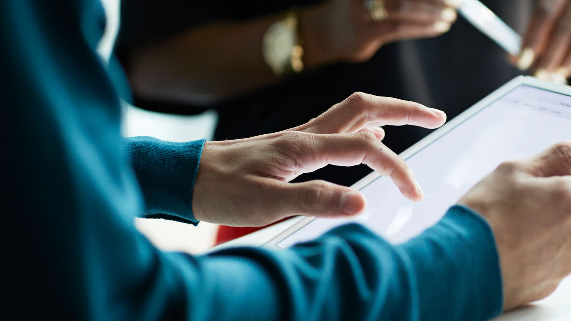 Close-up of someone using a tablet with a person holding a pen with both hands in the background