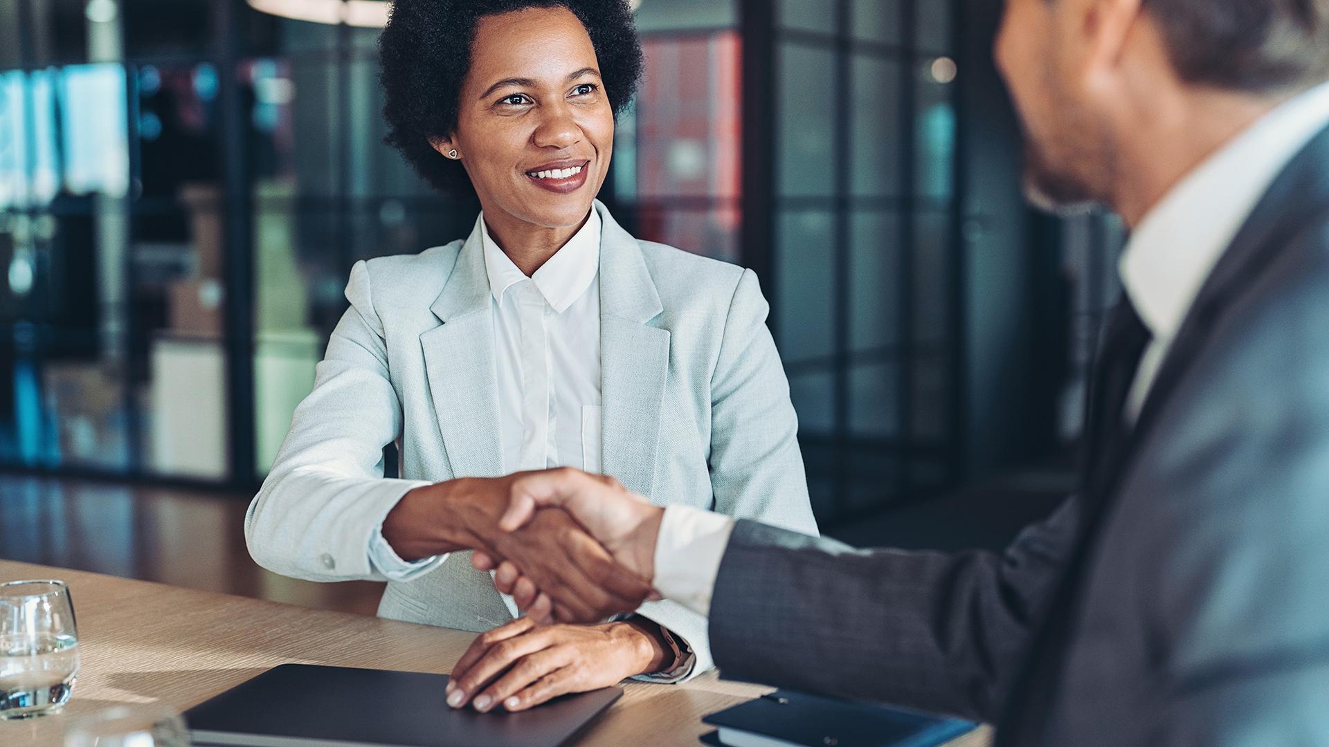Two people sitting across from each other while shaking hands at a desk