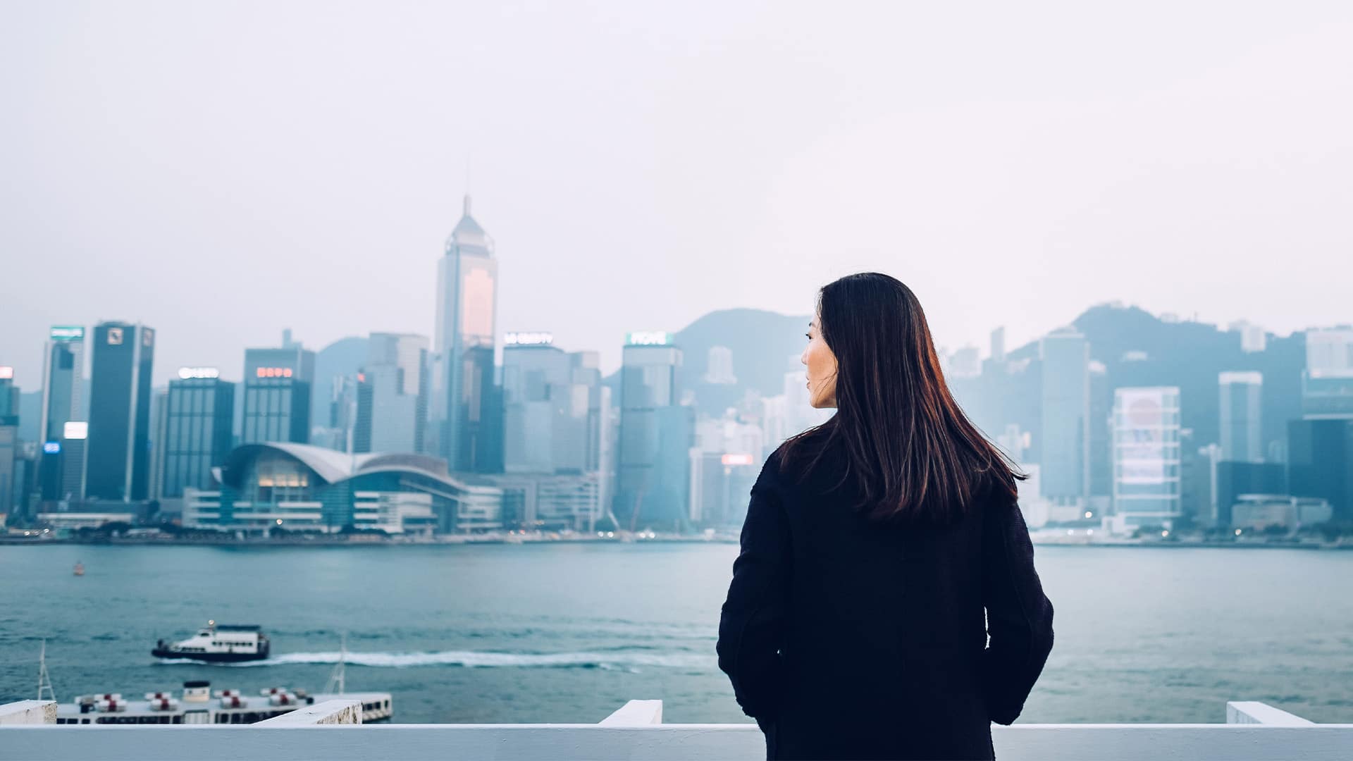 Rear view of young woman standing against the promenade of Victoria Harbour overlooking the cityscape of Hong Kong.