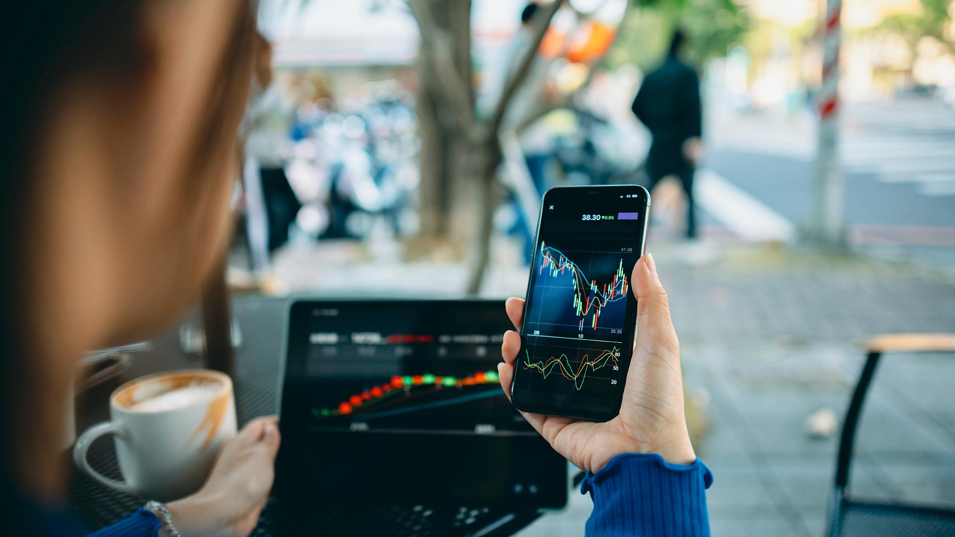 A person sitting outside a cafe using a smartphone and laptop to analyze data