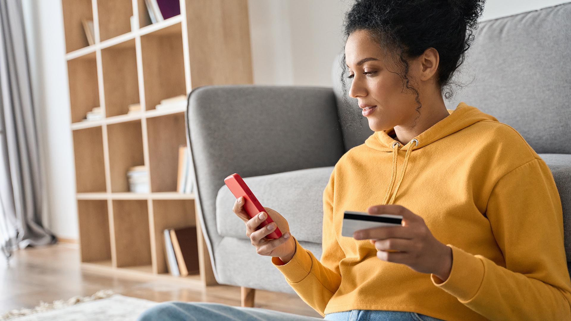 A women sitting in front of a couch while looking at and holding a smartphone and a credit card.