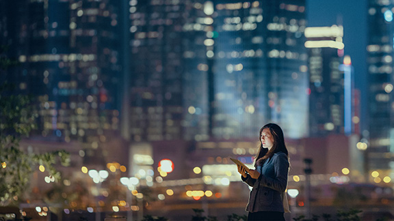 Young businesswoman using digital tablet in financial district, against illuminated corporate skyscrapers at night.