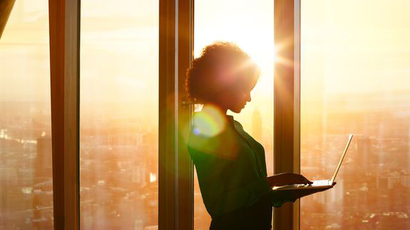 A women who is standing while holding and looking at a laptop with the sun rising in the background