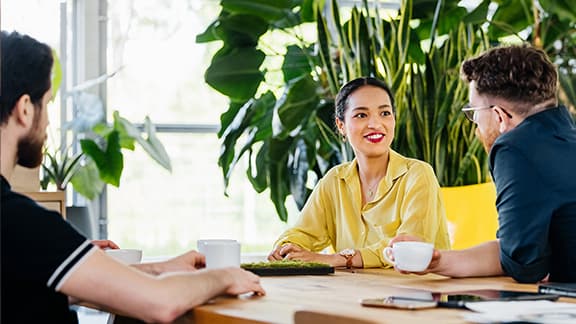 Three young professionals chatting around a large office desk with plants in the background.