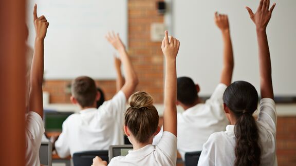 Students seated in a classroom raise their hands while facing a whiteboard.