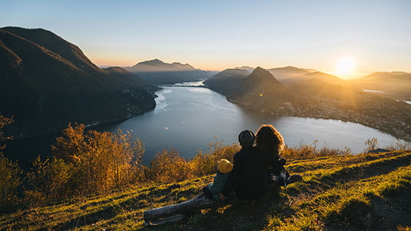A family sitting on a mountain top looking out at the lake, sunset, and other mountains