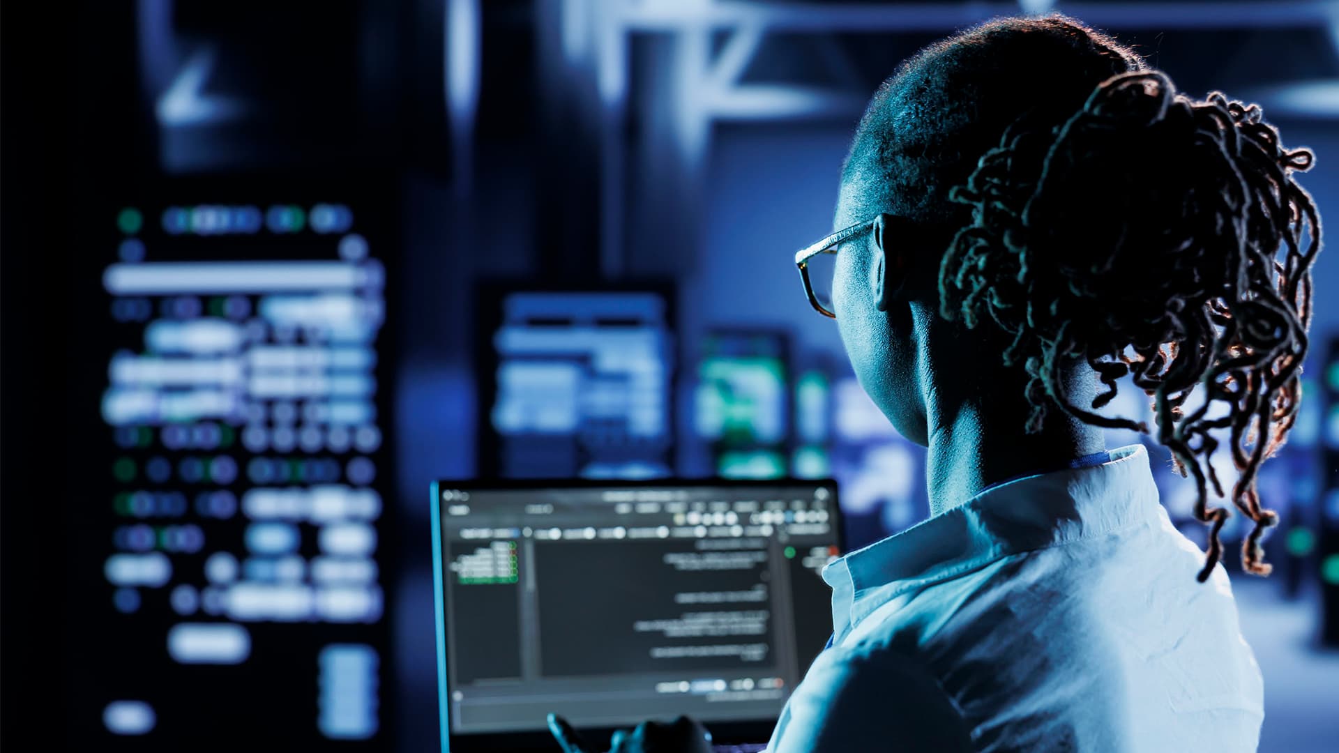 A person who is in an office surrounded by computers while the person is holding and working on a laptop