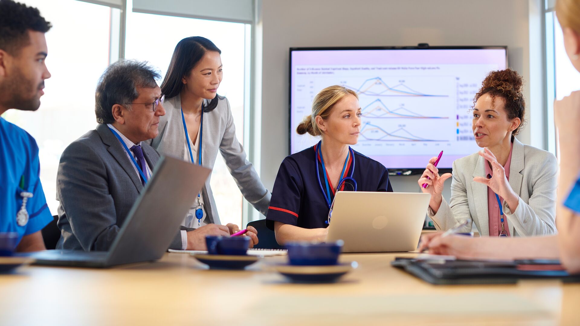 Healthcare team in a meeting discussing data on a screen while collaborating around a table with laptops.