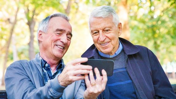 Two senior men, sitting in a park, looking at a video on an iphone.