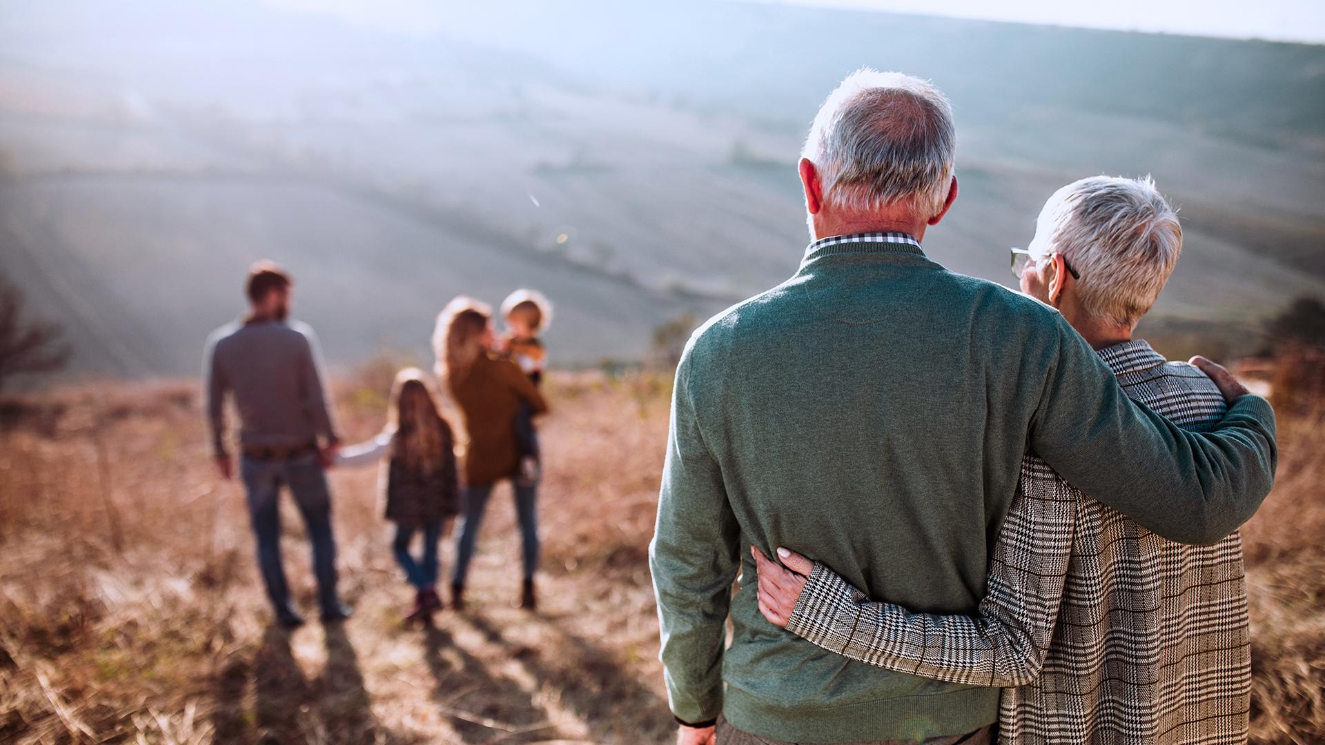 Back view of embraced grandparents enjoying while looking at their family on a field in autumn day.