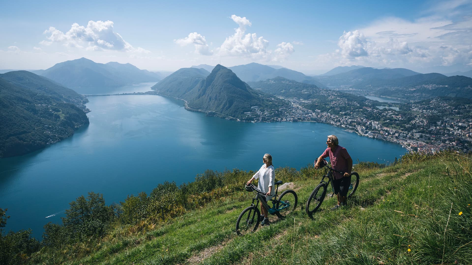 Two senior citizens pushing their bikes through a green valley bike trail.