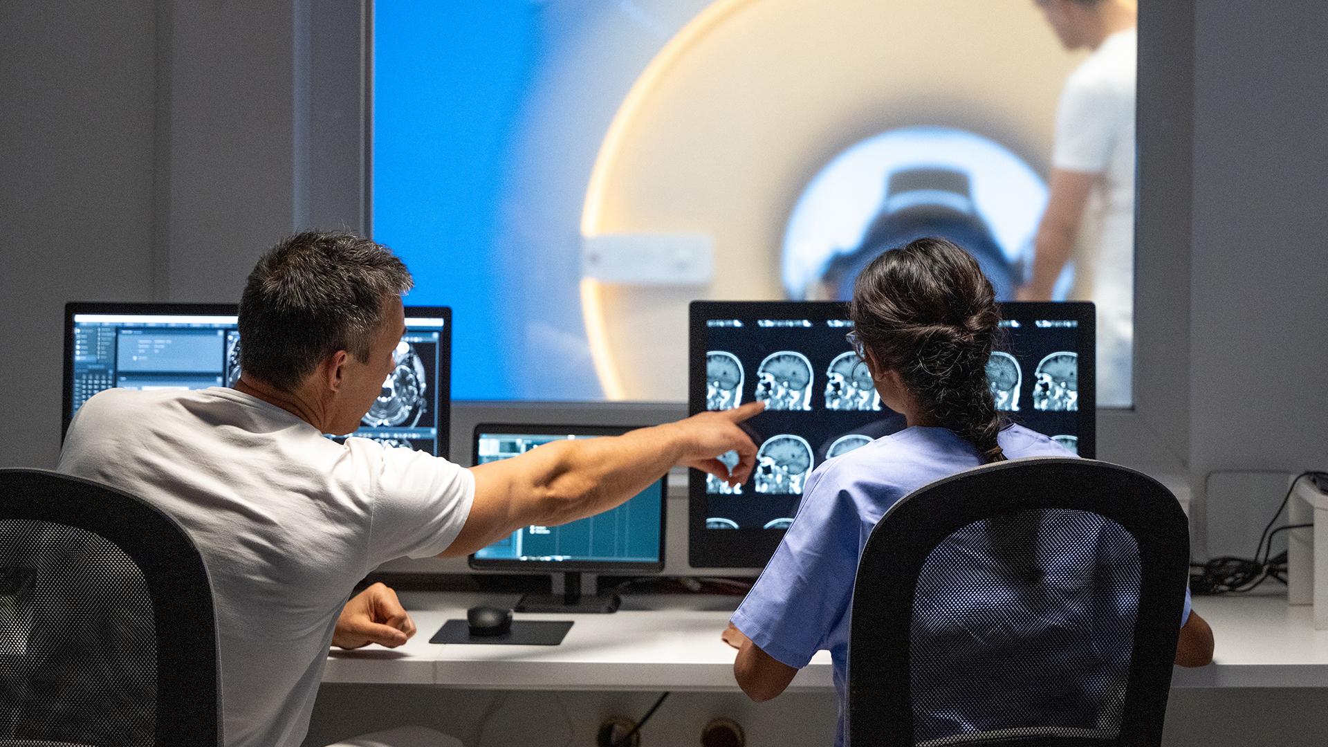 A male and female MRI technologists sitting at a desk in the operating room and operating the MRI scanner.