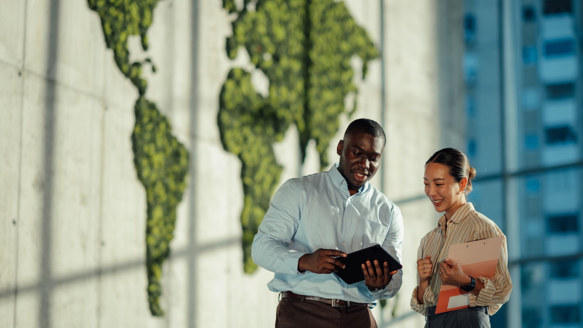 Two managers are discussing sustainable business strategies using digital tablet and clipboard, with a green world map.