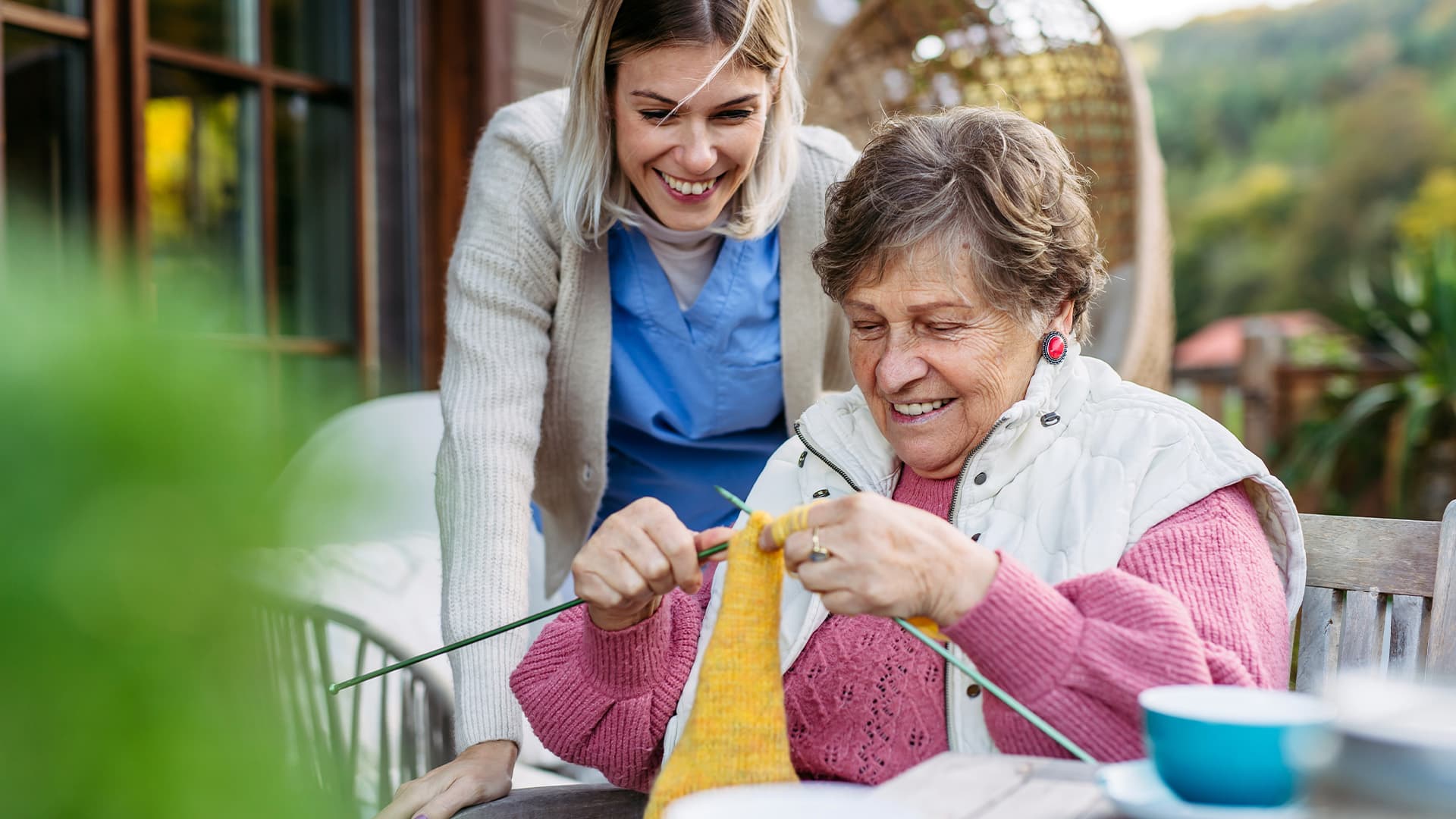 A senior woman knitting on an outdoor patio at a care facility.