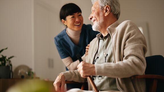 A smiling senior man sitting down and looking at his smiling female nurse.