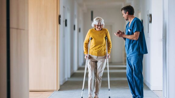 An elderly woman using crutches receives assistance from a nurse in a hospital corridor.
