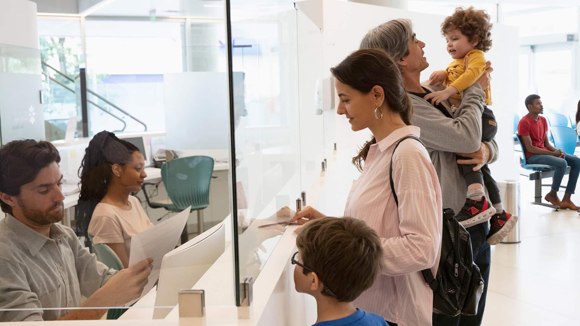 Family checks in at a medical reception desk while others sit in a waiting area behind glass.