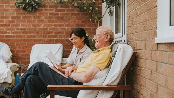 Female care assistant sits beside an elderly male and reads to him