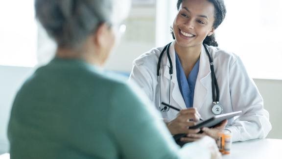 Doctor with a stethoscope smiling while speaking to an older patient and taking notes on a tablet.