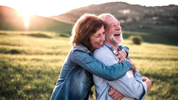 A senior couple who are smiling and hugging while outside in nature with the sun setting in the background