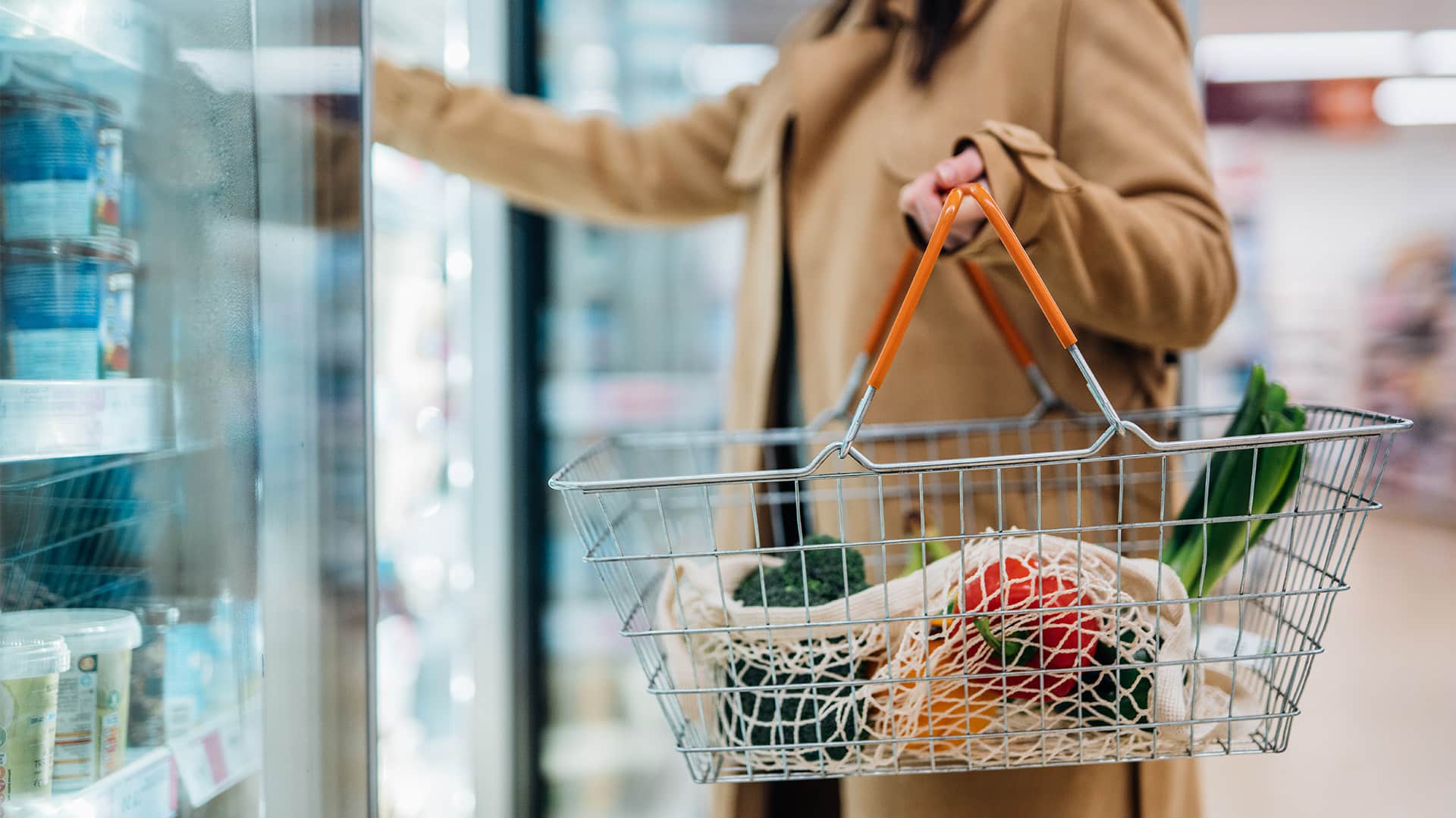 A woman looking in the dairy aisle at a grocery store while carrying a basket.