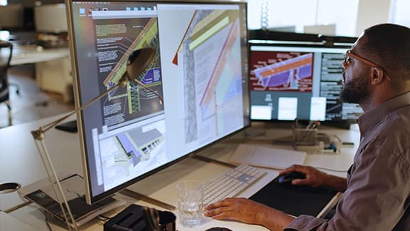 A man sitting at a desk while looking at a couple of computer monitors and going over construction plans