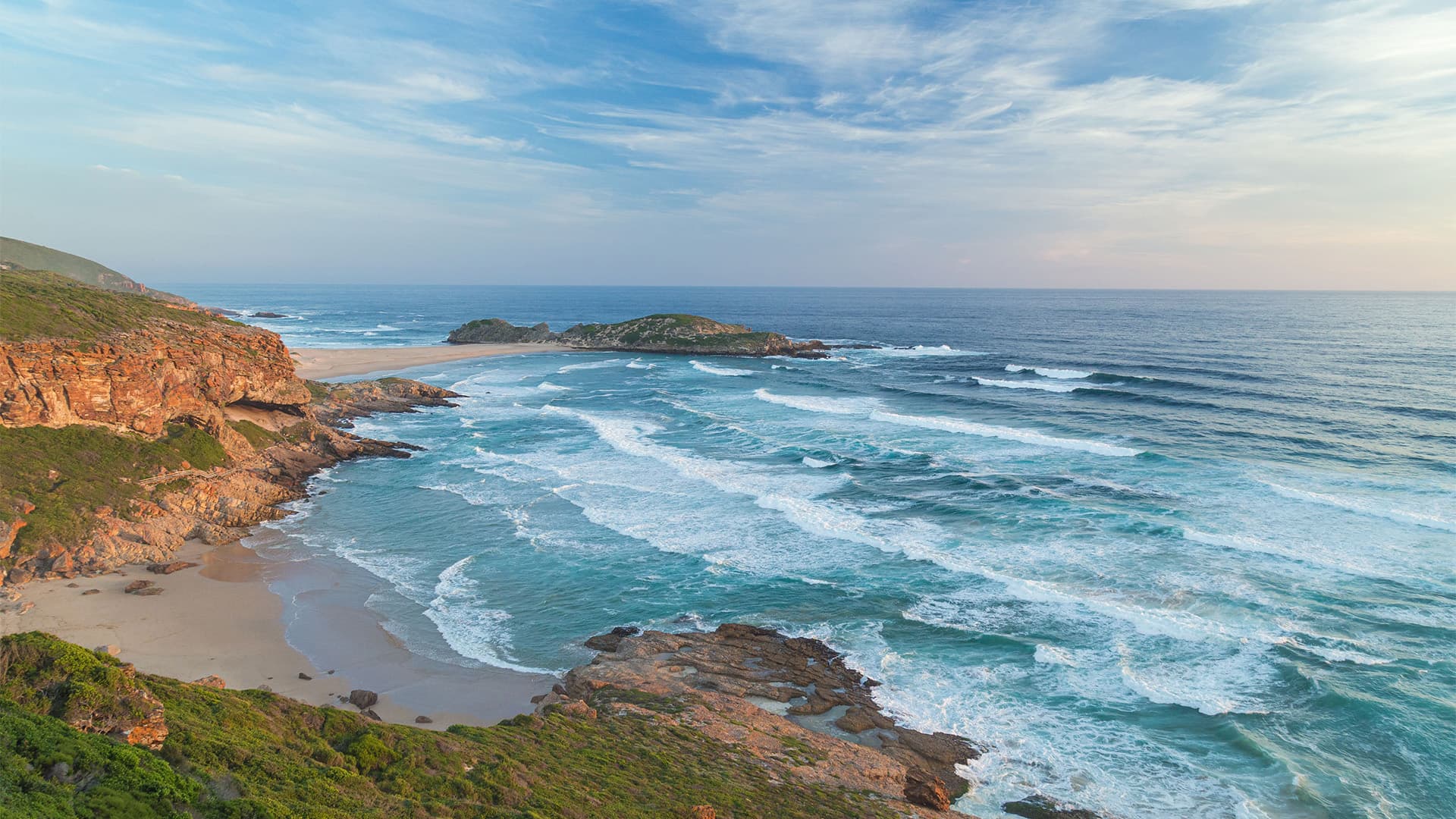 Rocky South African coastline with waves rolling onto a sandy cove under a blue, cloud‑streaked sky.
