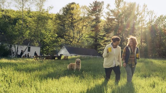 Two people walk through tall green grass in a sunlit field, with a dog nearby and trees and houses in the background.