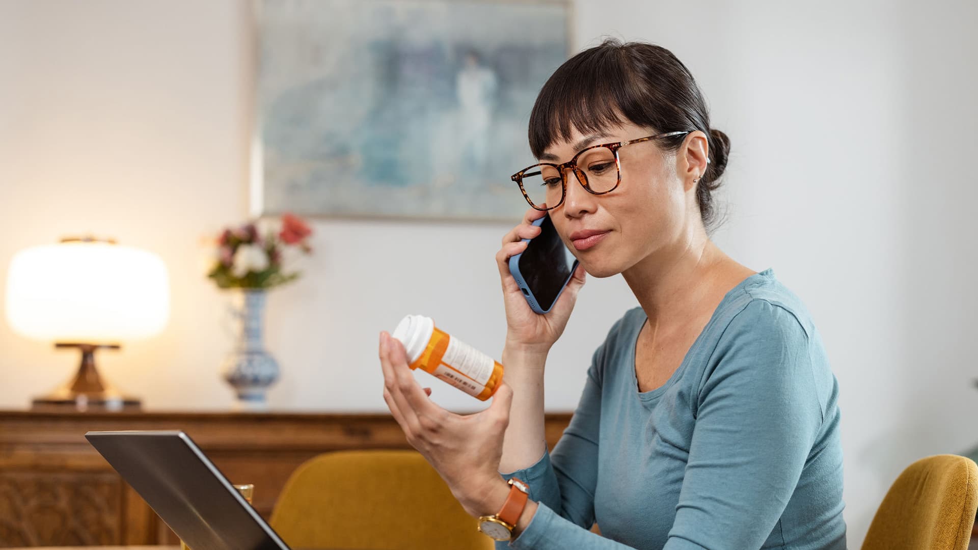 woman reading pill bottle instructions, talking on the phone and using laptop