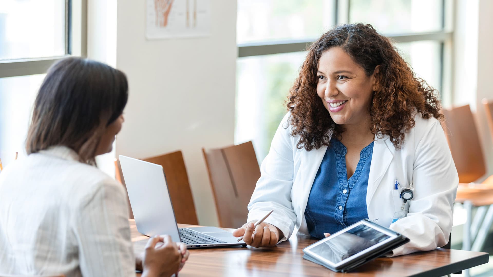 Healthcare professional in a white coat consulting with a patient at a table, with a laptop and tablet nearby.