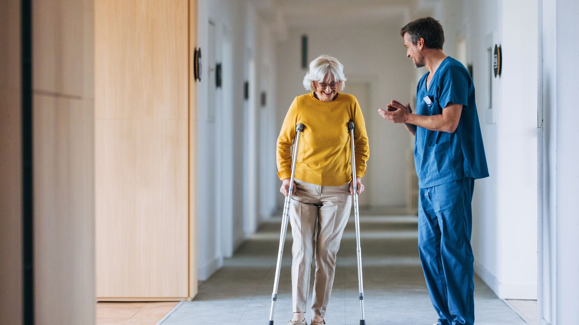An elderly woman using crutches receives assistance from a nurse in a hospital corridor.