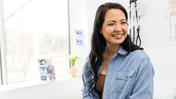 A patient sits on the examination table and smiles while listening to encouraging news from the doctor.