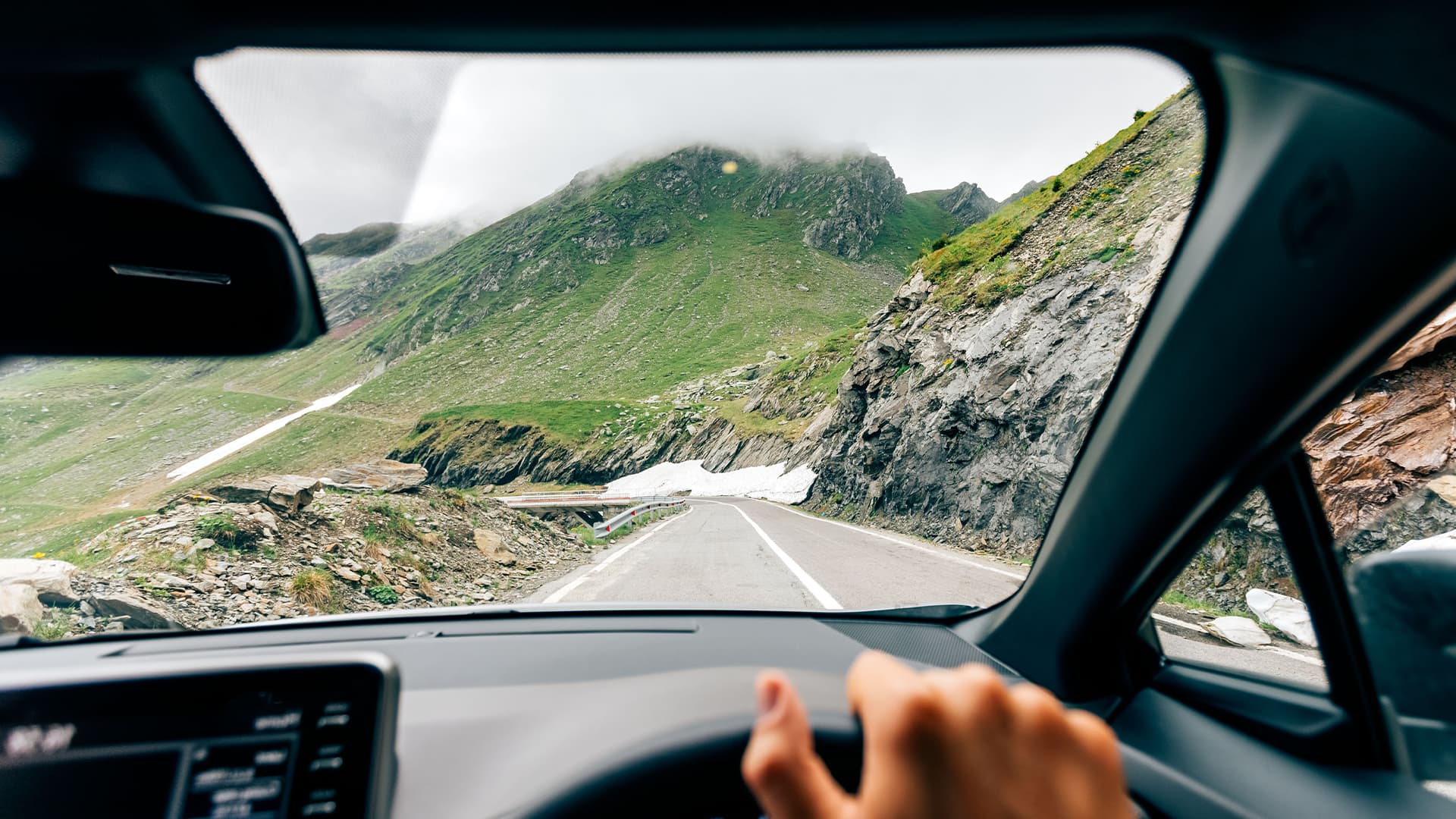 A person in a car driving down a mountain road with mountains, rocks, snow and greenery in the foreground
