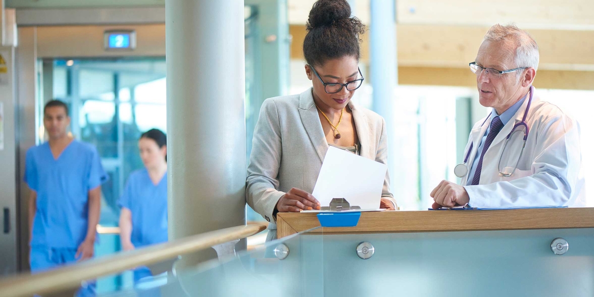A suited woman and a male doctor wearing a lab coat stand on a stairwell of a modern hospital and discuss some case notes.
