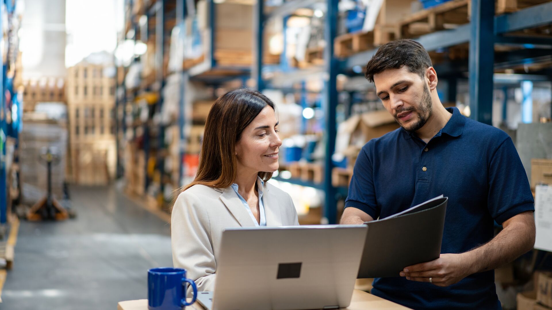 Woman manager and worker in a factory warehouse using a laptop to coordinate tasks and manage inventory.