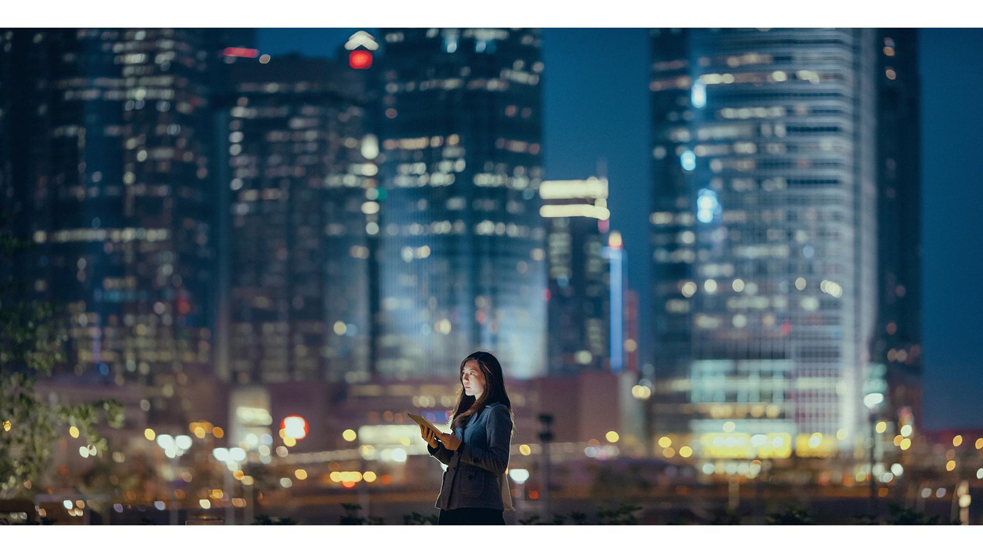 A women who is holding a digital tablet with buildings and skyscrapers in the background at nighttime