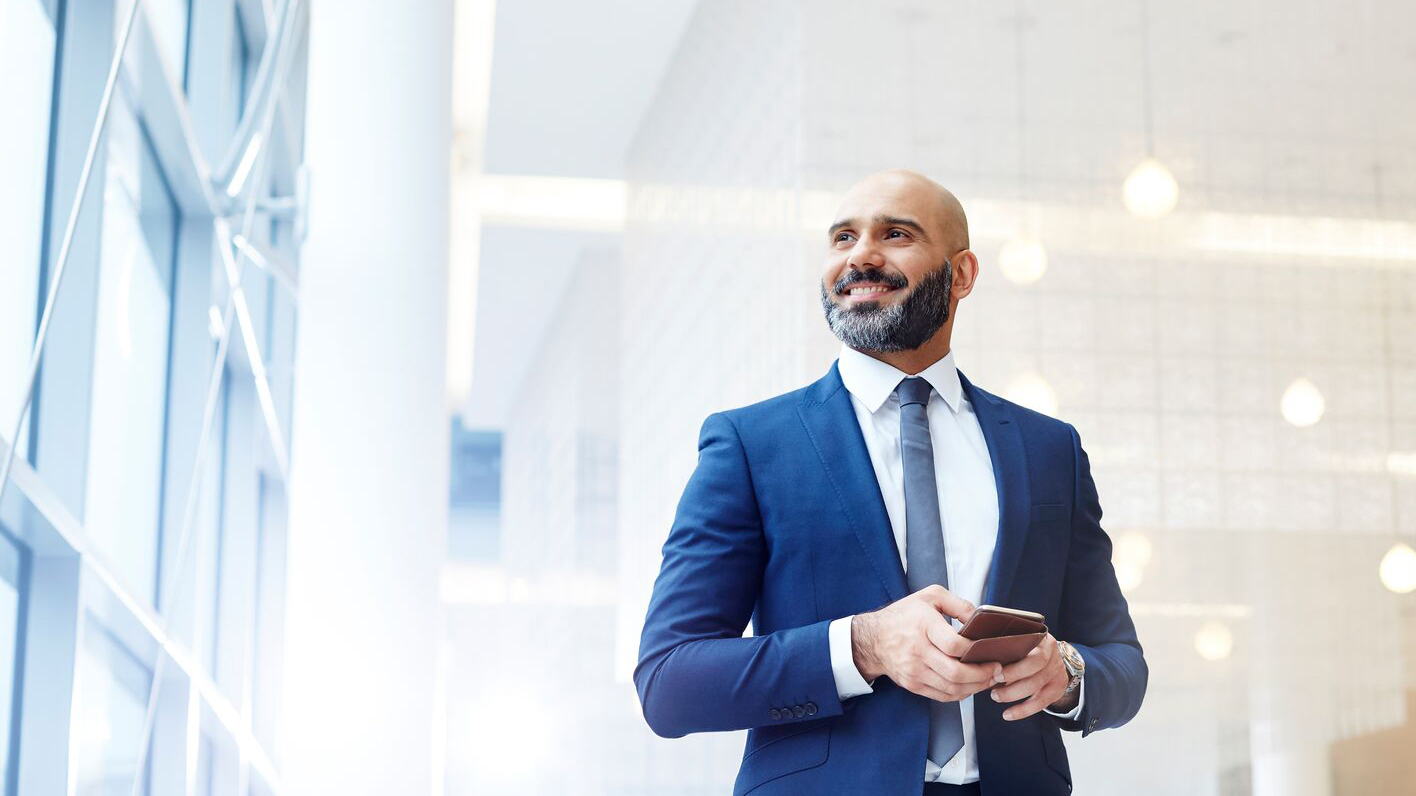 A well-dressed man in a suit stands in a bright lobby, smiling while holding a smartphone.