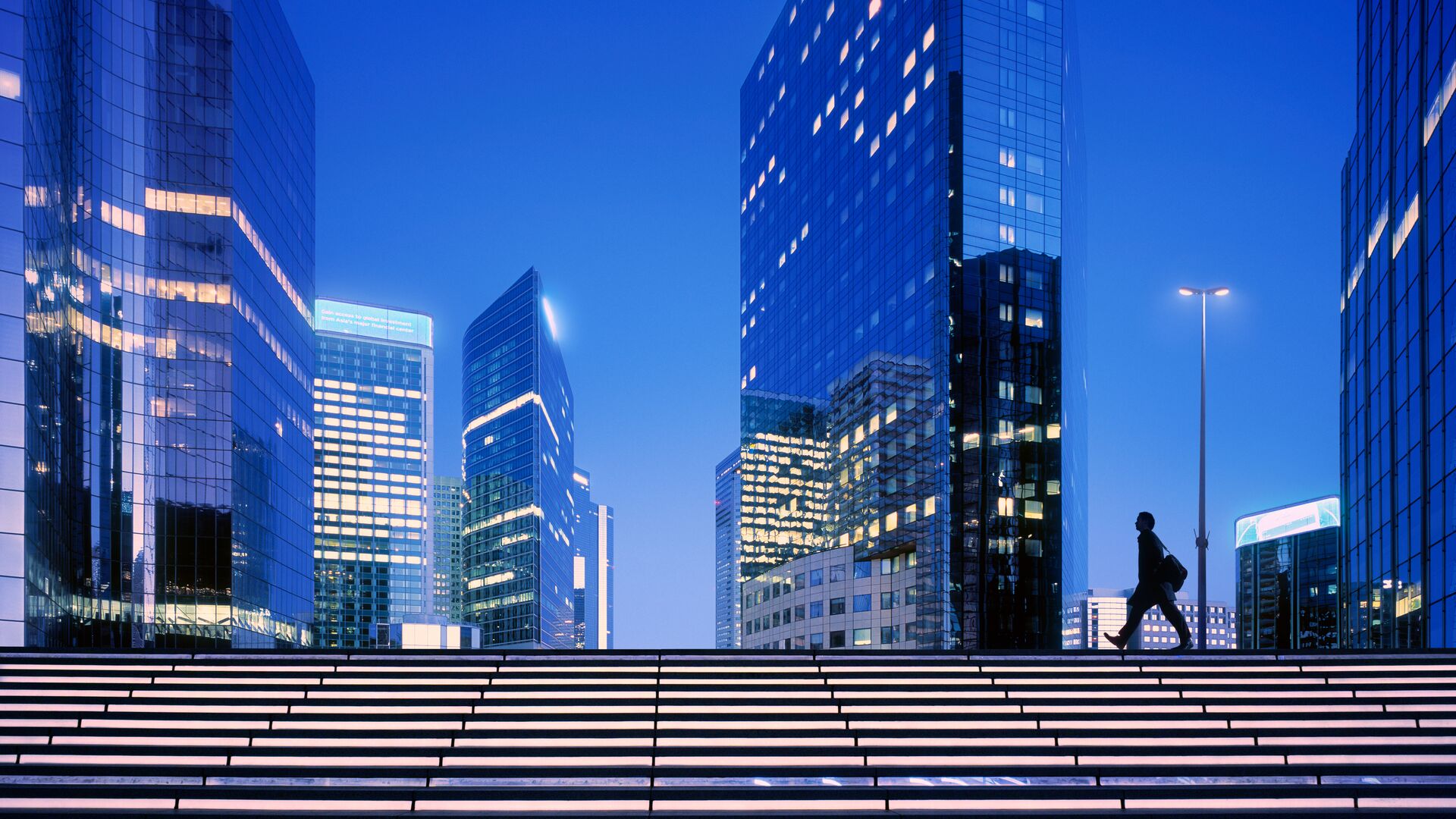 Modern city skyline at dusk with illuminated office towers, glowing steps, and a pedestrian crossing the plaza.