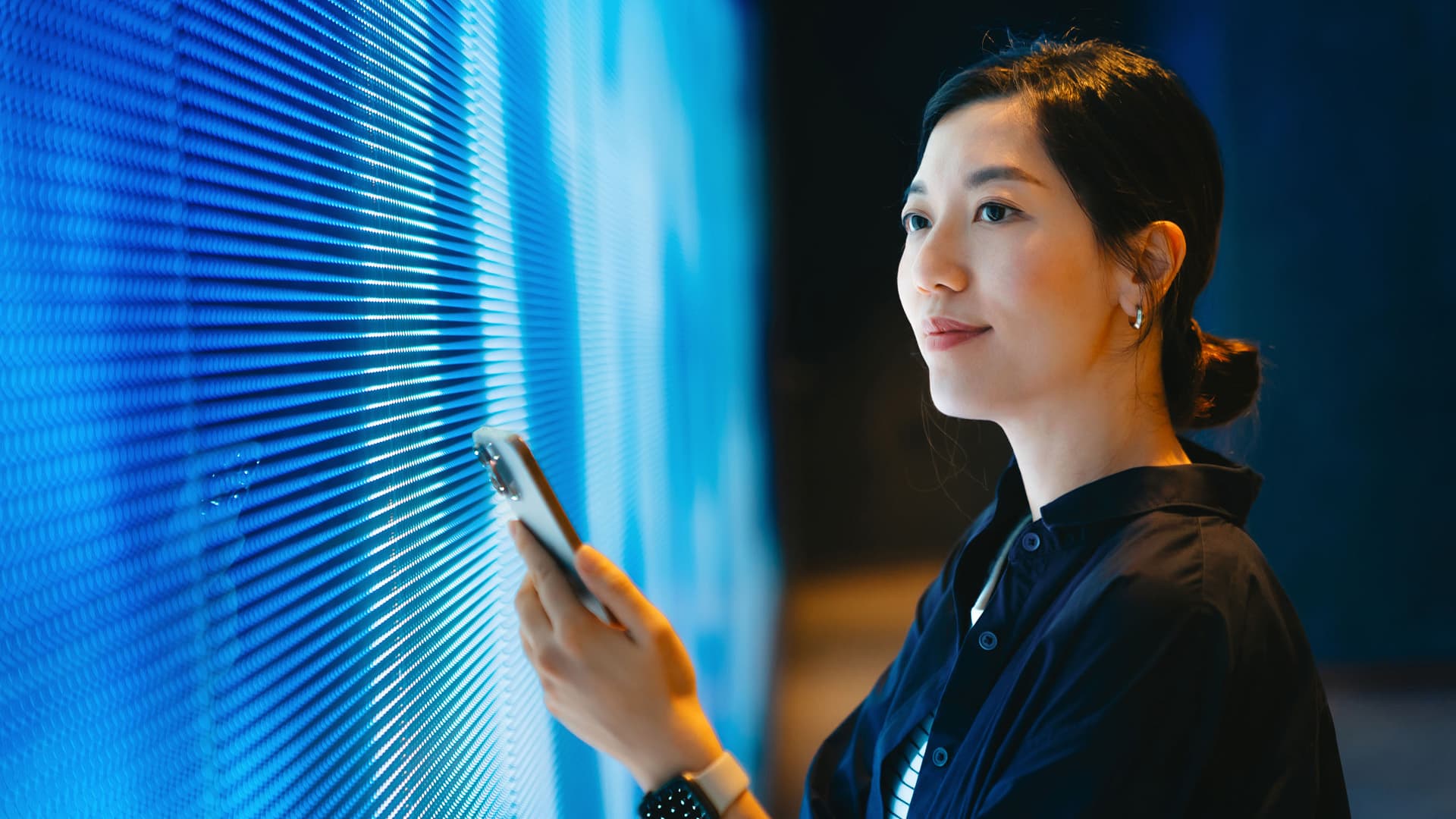 Confident young woman using smartphone against blue coloured illuminated LED digital display screen in the dark.
